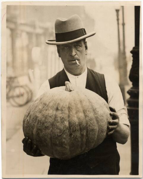 Man with hat and cigarette in mouth holding pumpkin at chest height