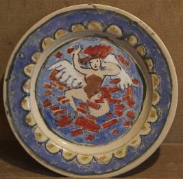 Angel with white wings and red rectangles around on blue plate with white scallops around rim