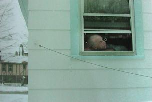 Head of figure in window looking up and right; window in light blue house with other house visible at lower left