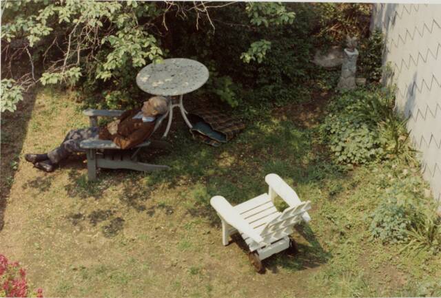 Man in lawn chair, seen from above, laying back next to white table under tree branches, with white chair at lower right