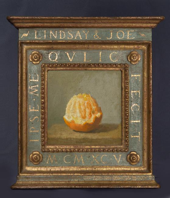 Painting of an orange, mostly peeled with rind remaining on bottom sitting on wood table in grey background. This painted image sits inside an ornate frame with gold borders, painted blue wiht the lettering "Lindsay & Joe" in gilded paint. A dark blue background surrounds the frame.
