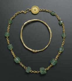 Gold necklace with emerald beans laid flat around gold bracelet sitting on black table