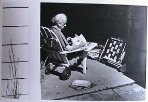 Man sitting in wooden lawn chair in garage, looking at boo, with plastic lawn chair at right